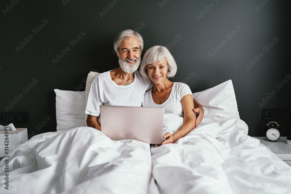 Happy family. Cozy home. Beautiful senior woman and her husband using laptop computer together in bedroom. Happy family. Cozy home. Beautiful senior woman and her husband using laptop computer together in bedroom.