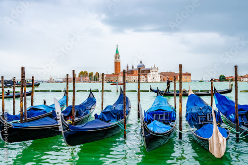 Venice, Italy. Gondolas/ Gondole docked by wooden mooring poles. Famous romantic tour boat ride for tourists/ couples/ people. Bell Tower of San Giorgio Maggiore Basilica Church in background. © Debbie Ann Powell