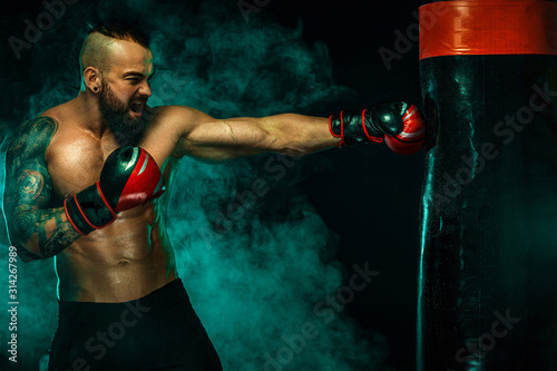 Boxer fighting in gloves with boxing punching bag. Sportsman with tattoos, man Isolated on black background with smoke. Copy Space. © Mike Orlov