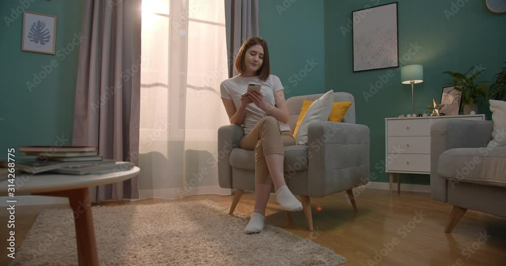 Young caucasian good looking smiled girl tapping and typing on the smartphone in the comfortable living room. Indoors. Young caucasian good looking smiled girl tapping and typing on the smartphone in the comfortable living room. Indoors.