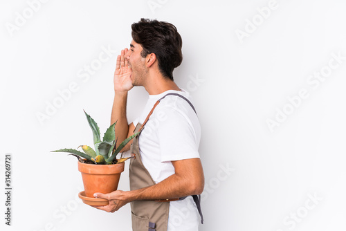 Young caucasian gardener man holding a plant isolated shouting and holding palm near opened mouth.