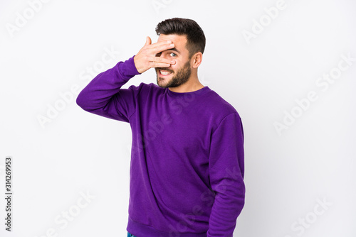 Young caucasian man against a white background isolated blink at the camera through fingers, embarrassed covering face.