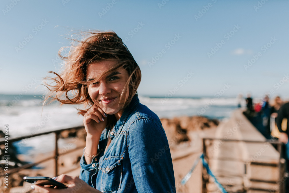 Excited lovable model enjoying outdoor photoshoot with smile. Happy woman listening music on the shore of ocean and posing to camera