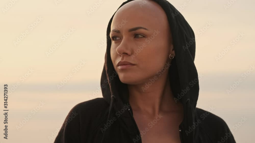 Smiling attractive bald in hood sports woman looking around while standing near the sea outdoors Smiling attractive bald in hood sports woman looking around while standing near the sea outdoors