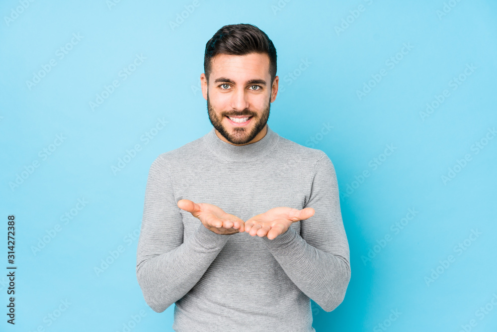 © Asier - Young caucasian man against a blue background isolated holding something with palms, offering to camera.