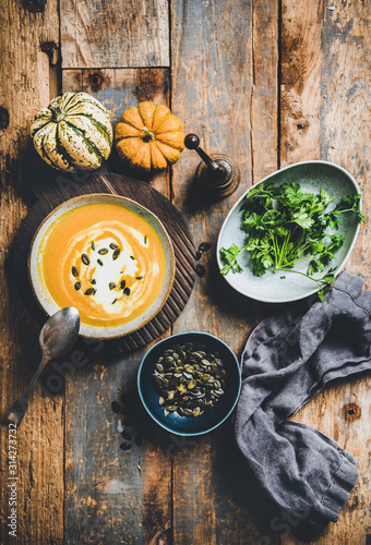 Autumn, winter warming seasonal meal. Flat-lay of pumpkin soup with seeds, fresh parsley and cream in bowl on board over wooden background, top view. Vegan, vegetarian, healthy, comfort food concept