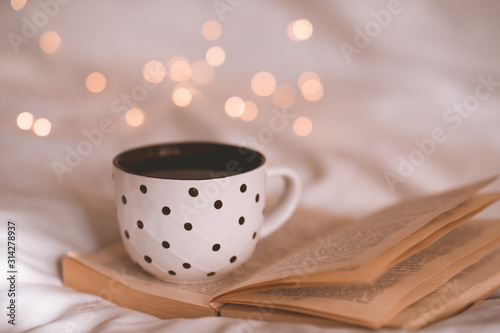 Open paper book with mug of coffee in bed over glowing lights close up. Good morning. Breakfast time.