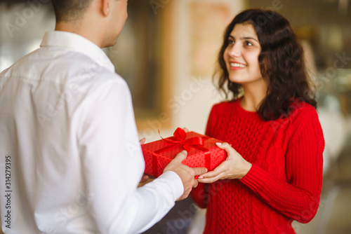 Man gives to his woman a gift box with red ribbon. A loving couple cuddles and celebrating Valentine's Day in the restaurant. Valentine's Day, holiday and surprise concept. Relationship and love. © maxbelchenko