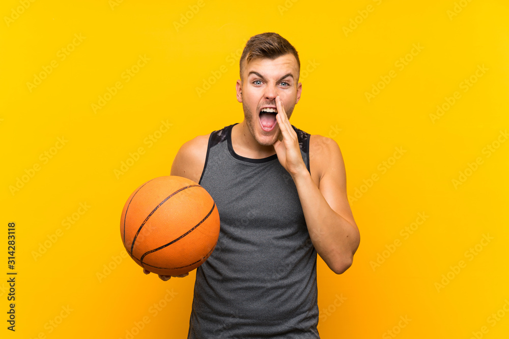 Young handsome blonde man holding a basket ball over isolated yellow background shouting with mouth wide open