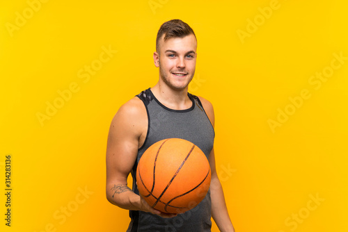 Young handsome blonde man holding a basket ball over isolated yellow background with happy expression