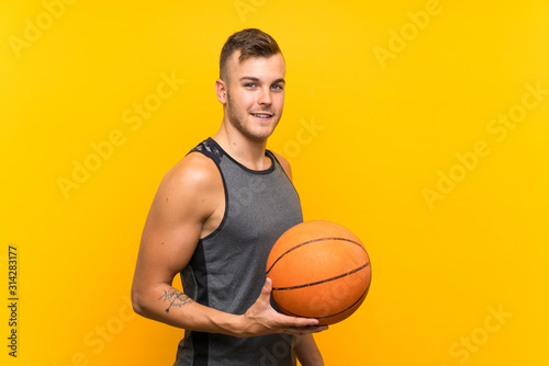 Young handsome blonde man holding a basket ball over isolated yellow background smiling a lot