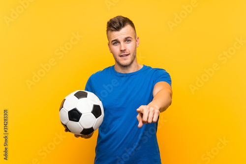 Young handsome blonde man holding a soccer ball over isolated yellow background points finger at you with a confident expression