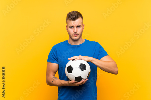 Young handsome blonde man holding a soccer ball over isolated yellow background