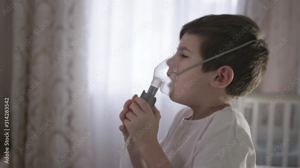 sick child inhaling through inhaler mask at home, treats inflammation of airways via nebulizer for preventing runny nose, cough, flu sitting on the bed sick child inhaling through inhaler mask at home, treats inflammation of airways via nebulizer for preventing runny nose, cough, flu sitting on the bed