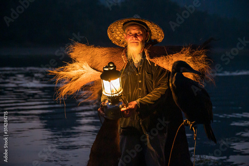 Fotografie An old fisherman hold a lamp on a raft with his cormorant at night