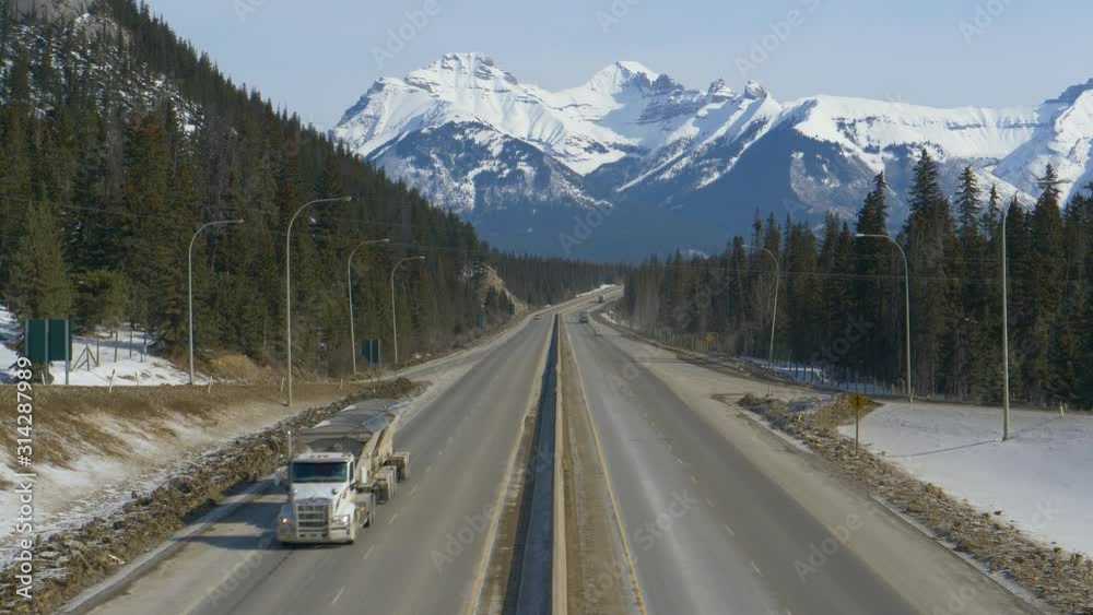 DRONE: Flying above the famous Trans Canada Highway as vehicles drive across the gorgeous wintry landscape. Cars and truck hauling cargo drive away from the stunning snowy mountain range in Alberta. DRONE: Flying above the famous Trans Canada Highway as vehicles drive across the gorgeous wintry landscape. Cars and truck hauling cargo drive away from the stunning snowy mountain range in Alberta.