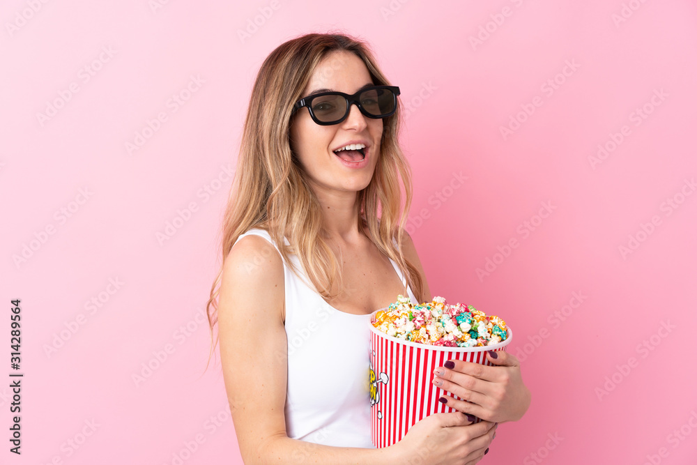 Young woman over isolated pink background with 3d glasses and holding a big bucket of popcorns