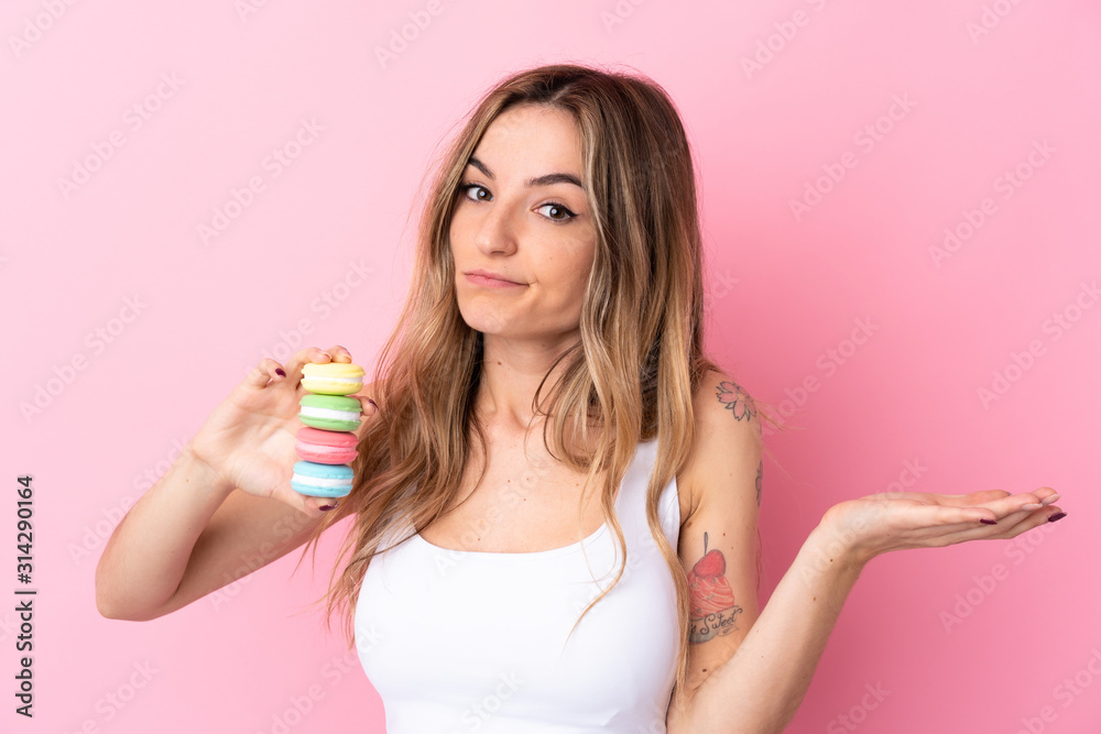 Young woman with  macarons over isolated pink background making doubts gesture while lifting the shoulders