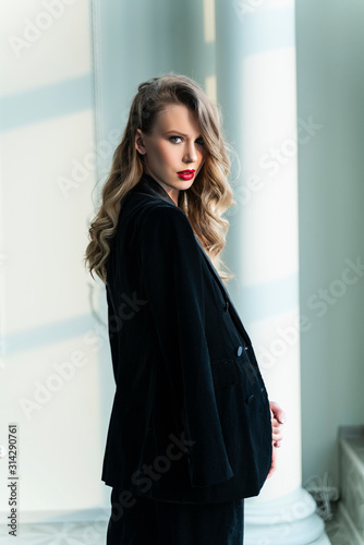 A beautiful slim blonde girl with red lips, wearing black pants and a fashionable blazer, stands at the window in a luxury vintage interior. Lifestyle, trendy and advertising design. © Алексей Торбеев