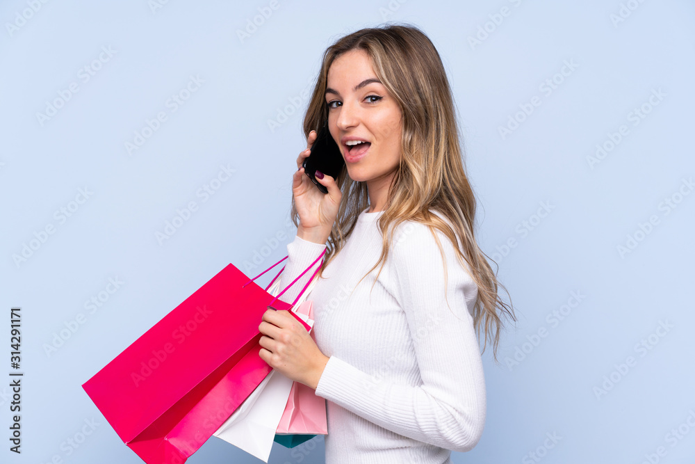 Young woman over isolated blue background holding shopping bags and calling a friend with her cell phone