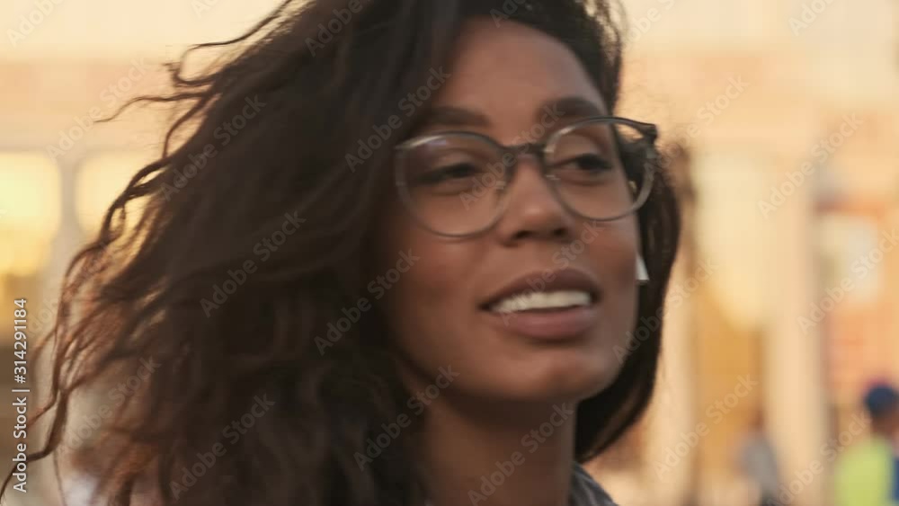 Close up view of Smiling pretty african woman in eyeglasses and earphones turning and going away outdoors Close up view of Smiling pretty african woman in eyeglasses and earphones turning and going away outdoors