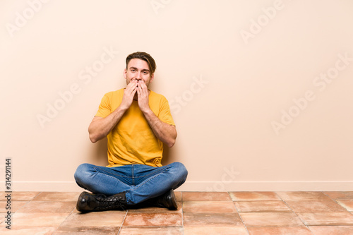 Young caucasian man sitting on the floor isolated laughing about something, covering mouth with hands.