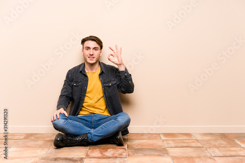 Young caucasian man sitting on the floor isolated winks an eye and holds an okay gesture with hand.