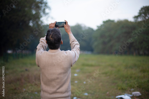 Back side portrait of an old Indian Bengali man in woolen sweater taking photograph of sky and trees by his cellphone in a field in winter afternoon in green background. Indian lifestyle and winter.