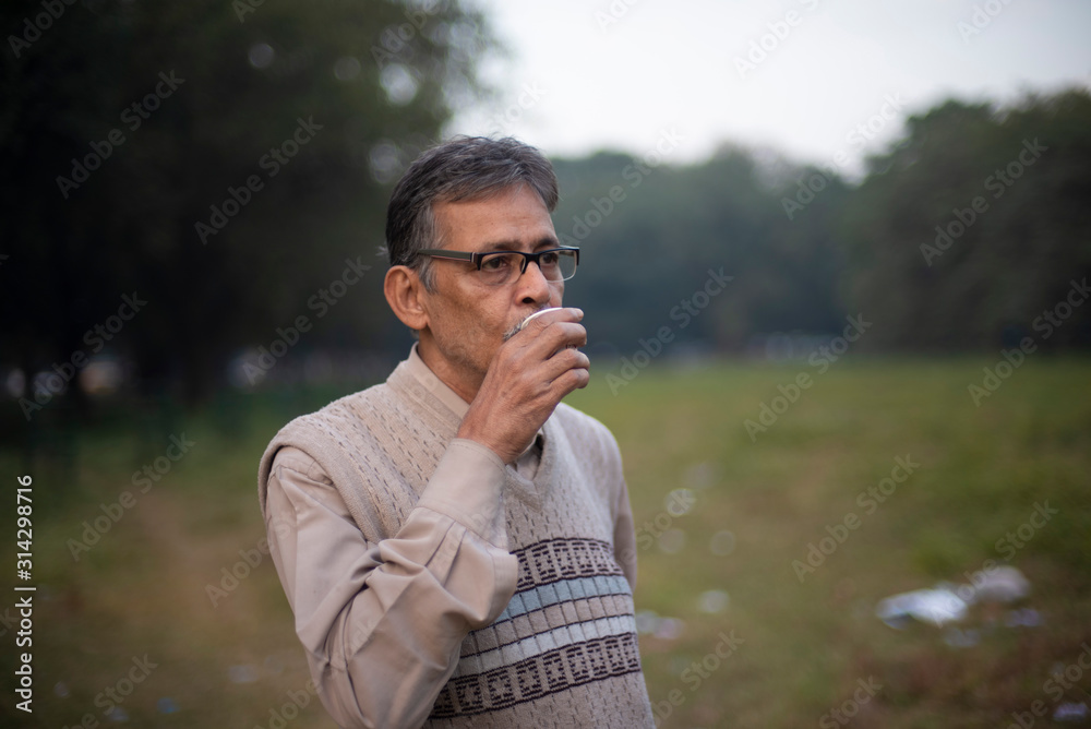 Portrait of an old Indian Bengali man in woolen sweater having tea/coffee in a field in winter afternoon in natural background. Indian lifestyle and winter. Portrait of an old Indian Bengali man in woolen sweater having tea/coffee in a field in winter afternoon in natural background. Indian lifestyle and winter.