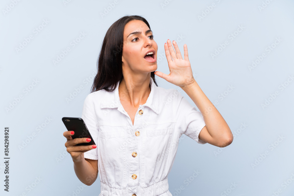 Young woman with a mobile phone over isolated blue wall shouting with mouth wide open Young woman with a mobile phone over isolated blue wall shouting with mouth wide open