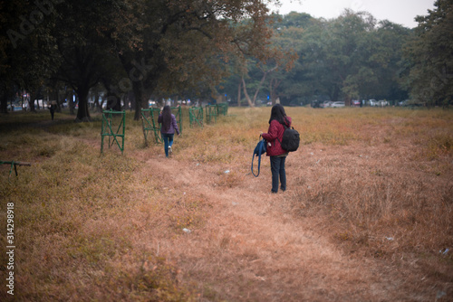 Back side of two young brunette Indian Bengali women/sisters with jackets and woolen sweater are walking in dry grass field in autumn afternoon in fall background. Indian lifestyle and fall.