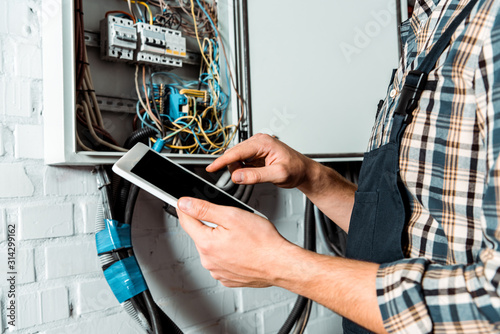 cropped view of electrician using digital tablet with blank screen near switchboard