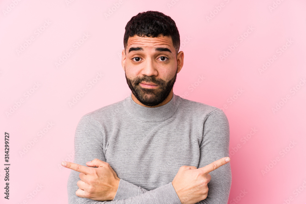 Young latin man against a pink background isolated points sideways, is trying to choose between two options. Young latin man against a pink background isolated points sideways, is trying to choose between two options.
