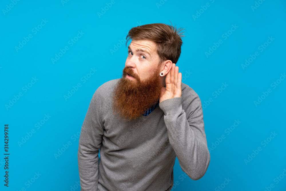Redhead man with long beard over isolated blue background listening to something by putting hand on the ear Redhead man with long beard over isolated blue background listening to something by putting hand on the ear
