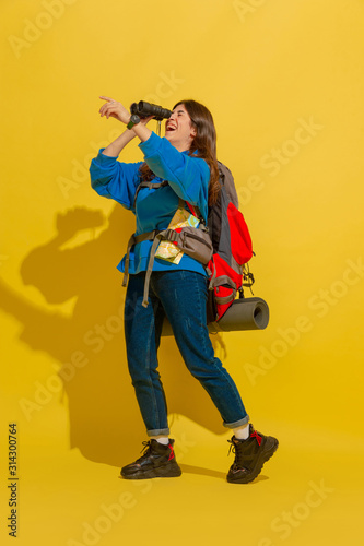 Smiling, looking up. Portrait of a cheerful young caucasian tourist girl with bag and binoculars isolated on yellow studio background. Preparing for traveling. Resort, human emotions, vacation.
