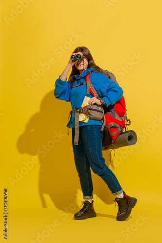 Smiling, looking up. Portrait of a cheerful young caucasian tourist girl with bag and binoculars isolated on yellow studio background. Preparing for traveling. Resort, human emotions, vacation.