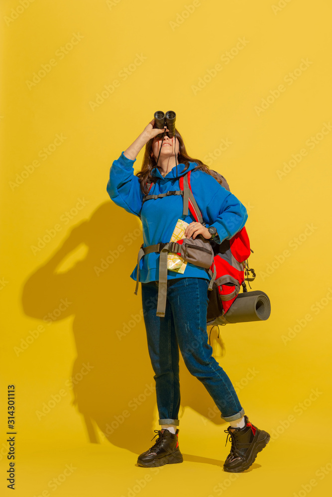 Smiling, looking up. Portrait of a cheerful young caucasian tourist girl with bag and binoculars isolated on yellow studio background. Preparing for traveling. Resort, human emotions, vacation. Smiling, looking up. Portrait of a cheerful young caucasian tourist girl with bag and binoculars isolated on yellow studio background. Preparing for traveling. Resort, human emotions, vacation.