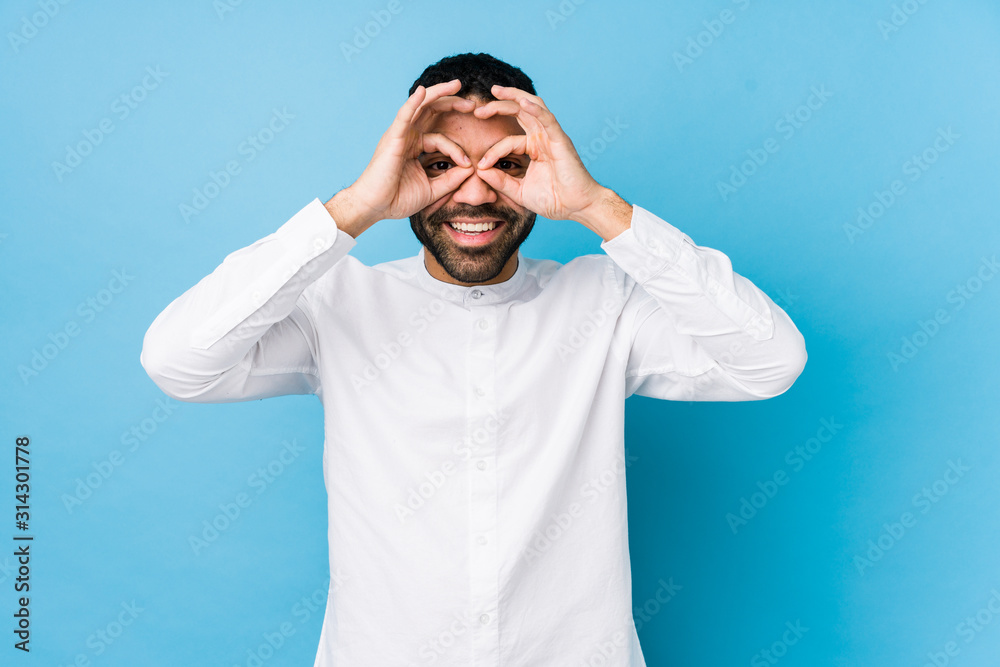 Young latin man against a blue background isolated showing okay sign over eyes Young latin man against a blue background isolated showing okay sign over eyes