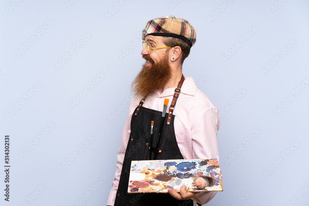 Redhead artist man with long beard holding a palette over isolated blue background with happy expression Redhead artist man with long beard holding a palette over isolated blue background with happy expression