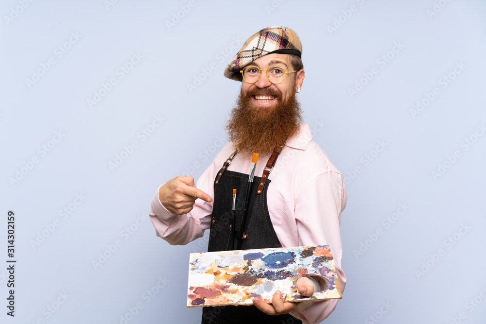 Redhead artist man with long beard holding a palette over isolated blue background and pointing it Redhead artist man with long beard holding a palette over isolated blue background and pointing it