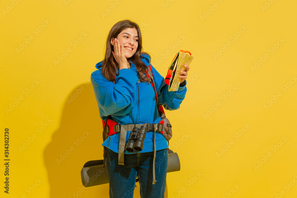 Smiling, looks dreamful. Portrait of a cheerful young caucasian tourist girl with bag and binoculars isolated on yellow studio background. Preparing for traveling. Resort, human emotions, vacation. Smiling, looks dreamful. Portrait of a cheerful young caucasian tourist girl with bag and binoculars isolated on yellow studio background. Preparing for traveling. Resort, human emotions, vacation.