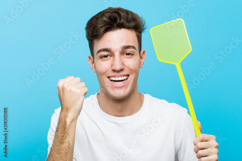 Young caucasian man holding a fly swatter cheering carefree and excited. Victory concept.