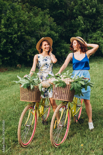 Two pretty girls go with bicycles in spring park at sunset. Girlfriends have fun, laugh, smile and ride bicycles. Full-length portrait of two young women in dresses and straw hats. active holiday