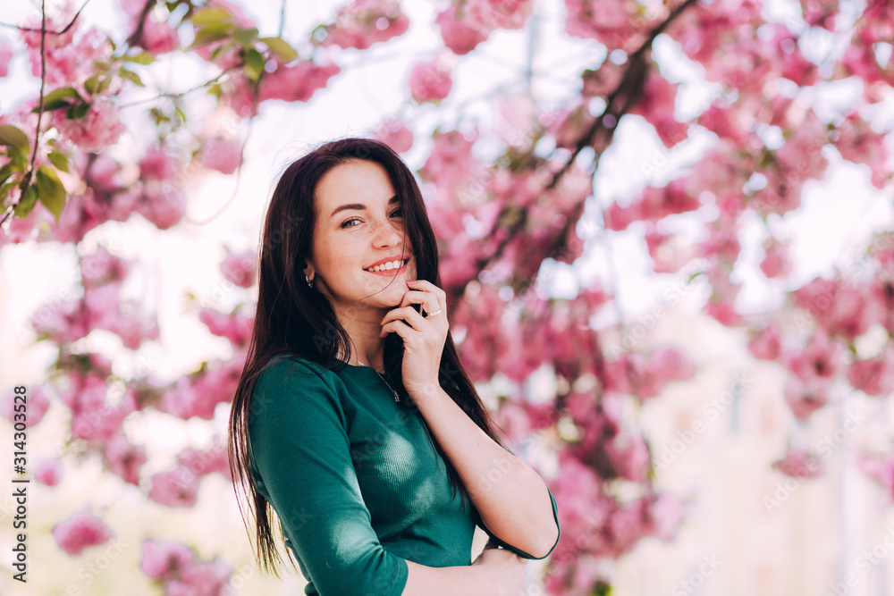 Closeup portrait of pretty girl in park with blooming japanese sakura trees. Romantic young brunette in green dress posing on background of spring flowering trees. Cherry blossoms. smiling girl. Closeup portrait of pretty girl in park with blooming japanese sakura trees. Romantic young brunette in green dress posing on background of spring flowering trees. Cherry blossoms. smiling girl.