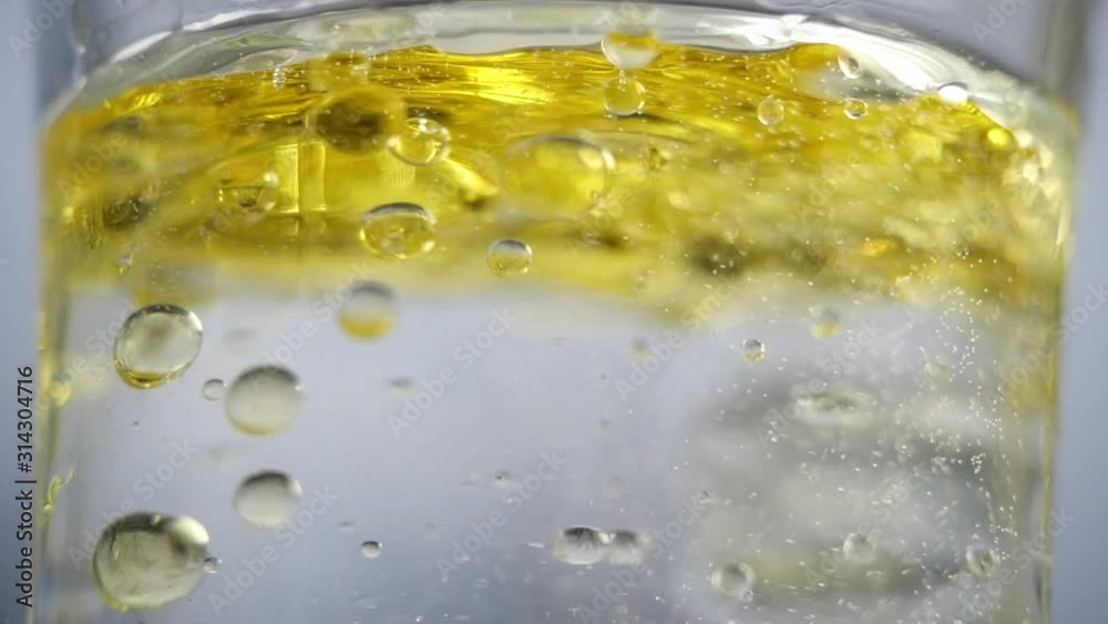 Beautiful shot near the liquid in a glass of water. Yellow oil is poured into a jar of clear water and bubbles with bulbs appear. Close view of a container of water that fills with a yellow liquid.