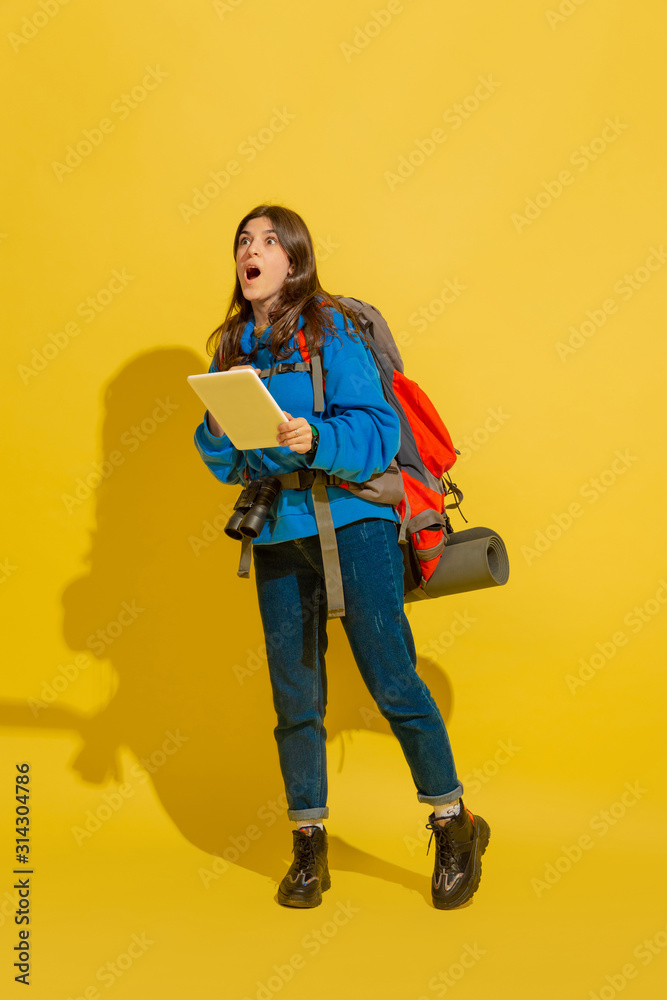 Looking for way with map. Portrait of a cheerful young caucasian tourist girl with bag and binoculars isolated on yellow studio background. Preparing for traveling. Resort, human emotions, vacation. Looking for way with map. Portrait of a cheerful young caucasian tourist girl with bag and binoculars isolated on yellow studio background. Preparing for traveling. Resort, human emotions, vacation.