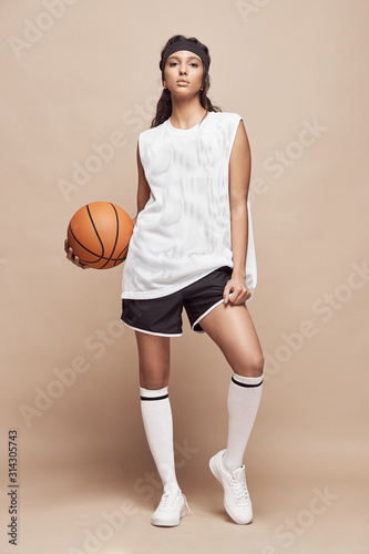 beautiful dark-skinned girl stands in full height on a beige background in white sports shirt, black shorts, white high socks and sneakers with a basketball ball and looks into the camera