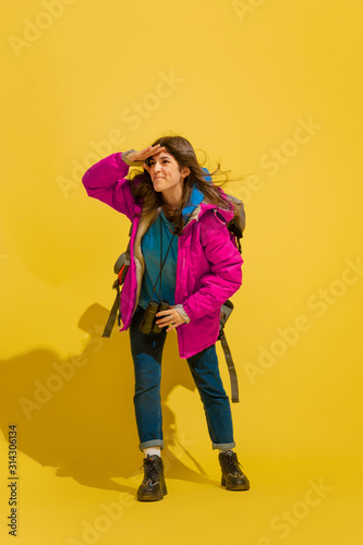 Smiling, looks happy. Portrait of a cheerful young caucasian tourist girl with bag and binoculars isolated on yellow studio background. Preparing for traveling. Resort, human emotions, vacation.