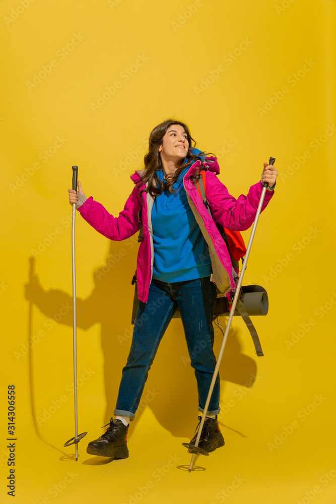 Going to mountains. Portrait of a cheerful young caucasian tourist girl with bag and binoculars isolated on yellow studio background. Preparing for traveling. Resort, human emotions, vacation.