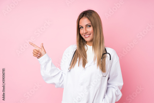 Young blonde woman over isolated pink background with doctor gown and pointing side